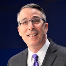 John Rademacher wearing glasses, a dark suit and a purple tie