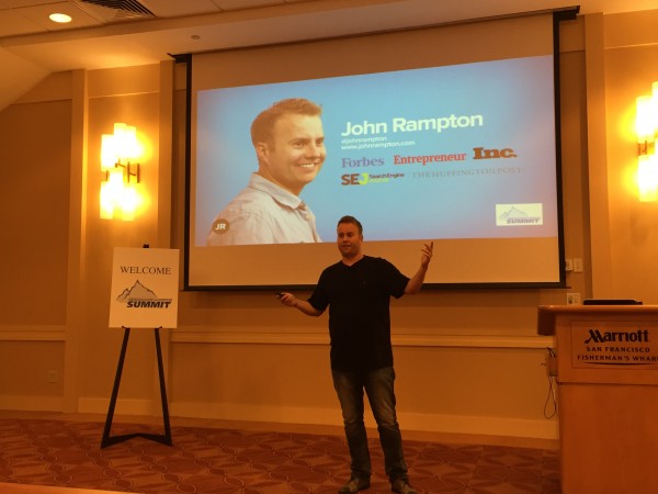 John Rampton speaking on a conference stage, arms outstretched, with his name projected behind him