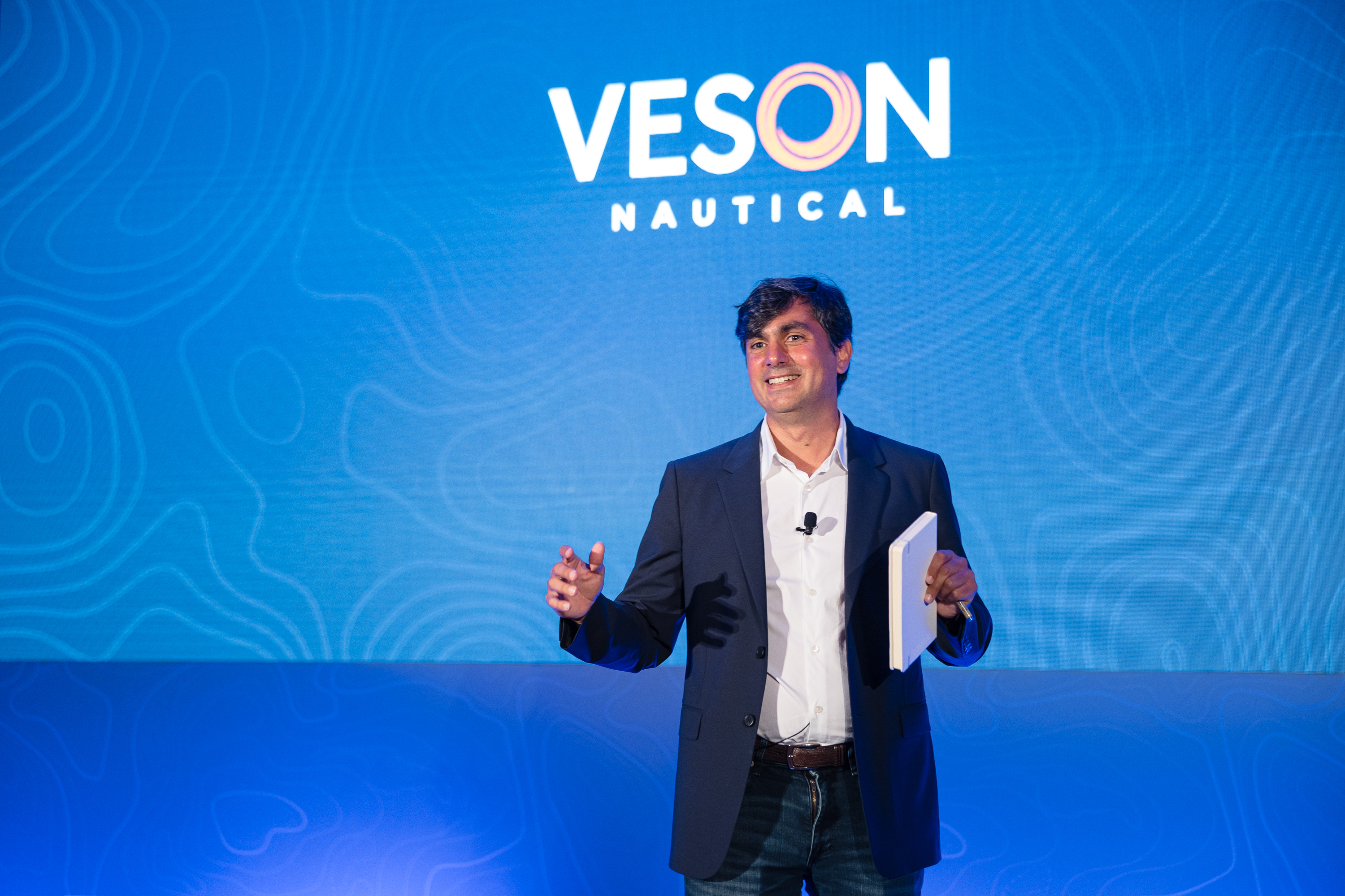John Veson speaking on stage, holding notes, against the blue Veson Nautical conference backdrop