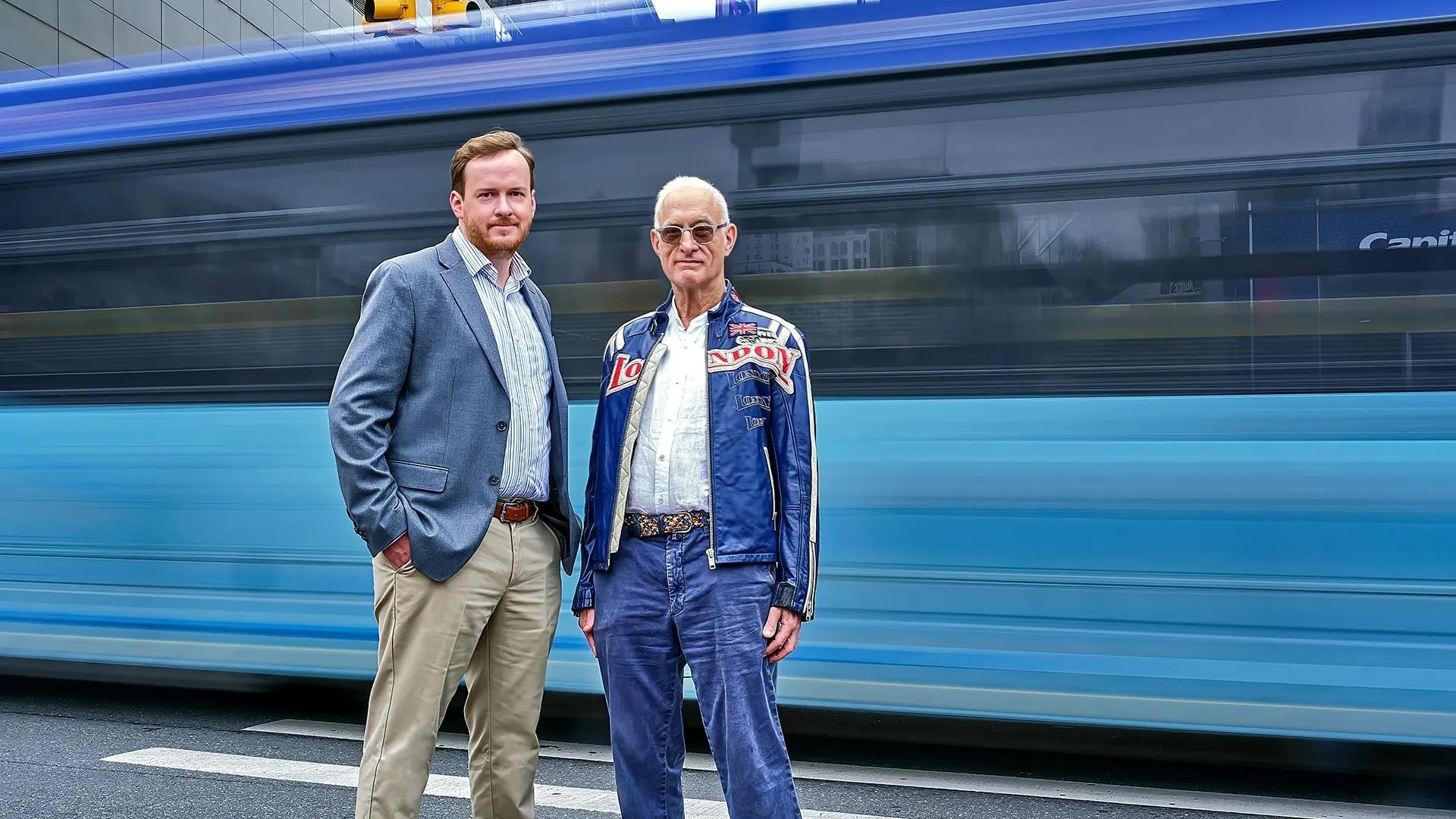 Andrew Kruegel, left, and Jonathan Sporn standing on a city street as a blue bus passes behind them