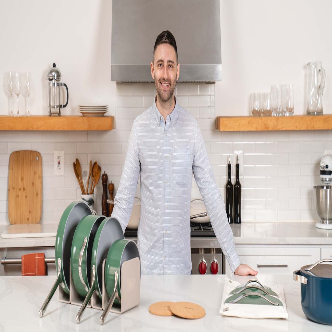 Jordan Nathan standing in a bright kitchen behind green Caraway cookware arranged in storage racks