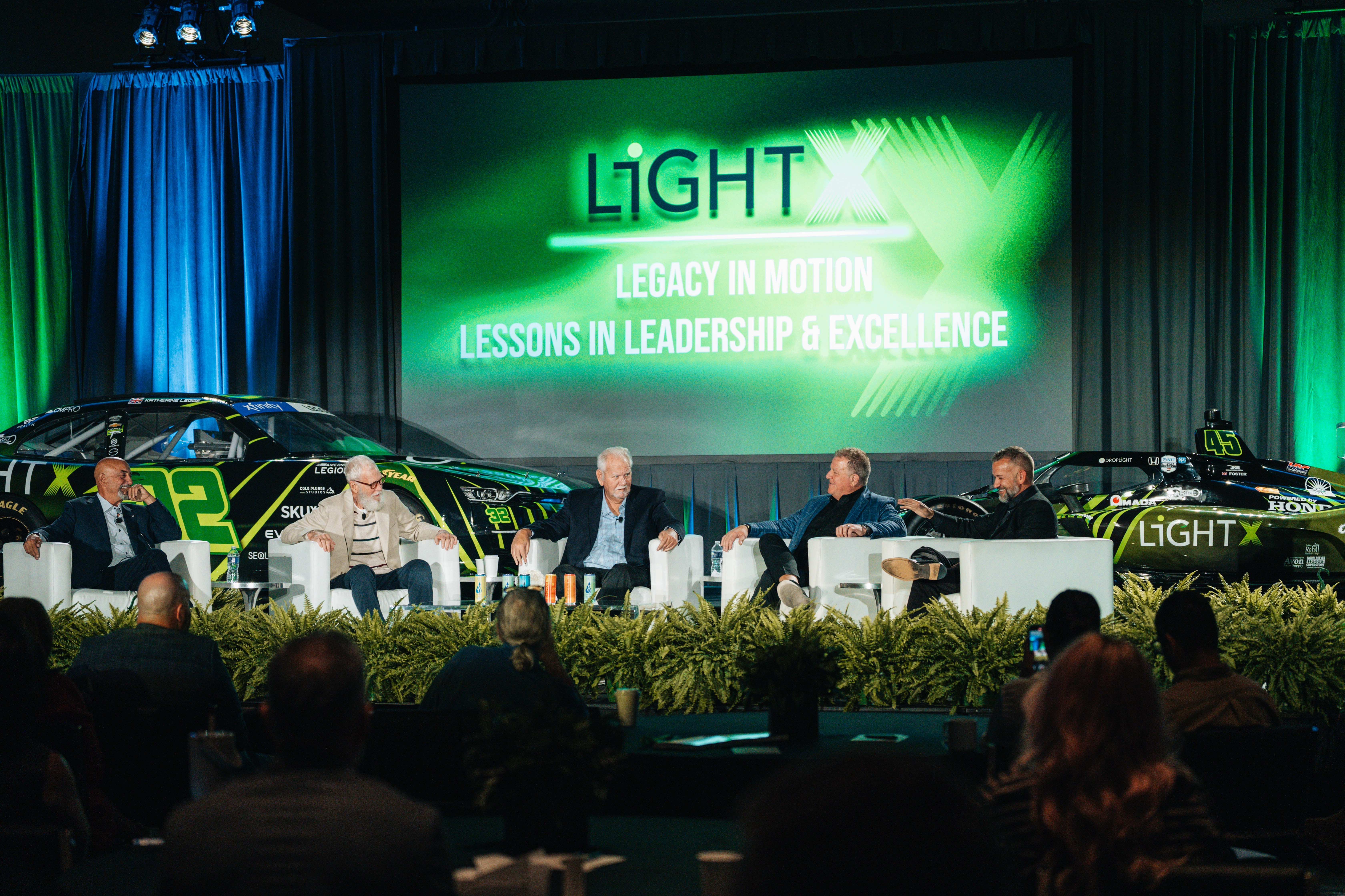Joseph Anderson at the far right moderating a LightX panel with four guests between two race cars.
