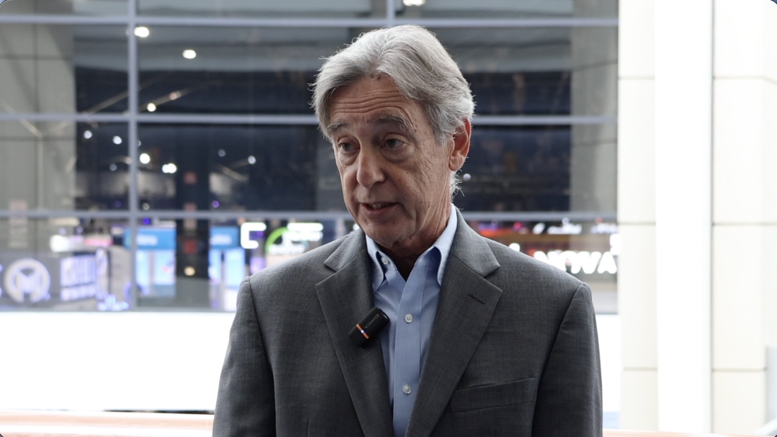 Joseph Belanoff wearing a gray jacket and speaking with an interviewer in a bright conference setting