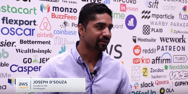 Joseph D'Souza speaking during an AWS Startups interview, with startup company names behind him