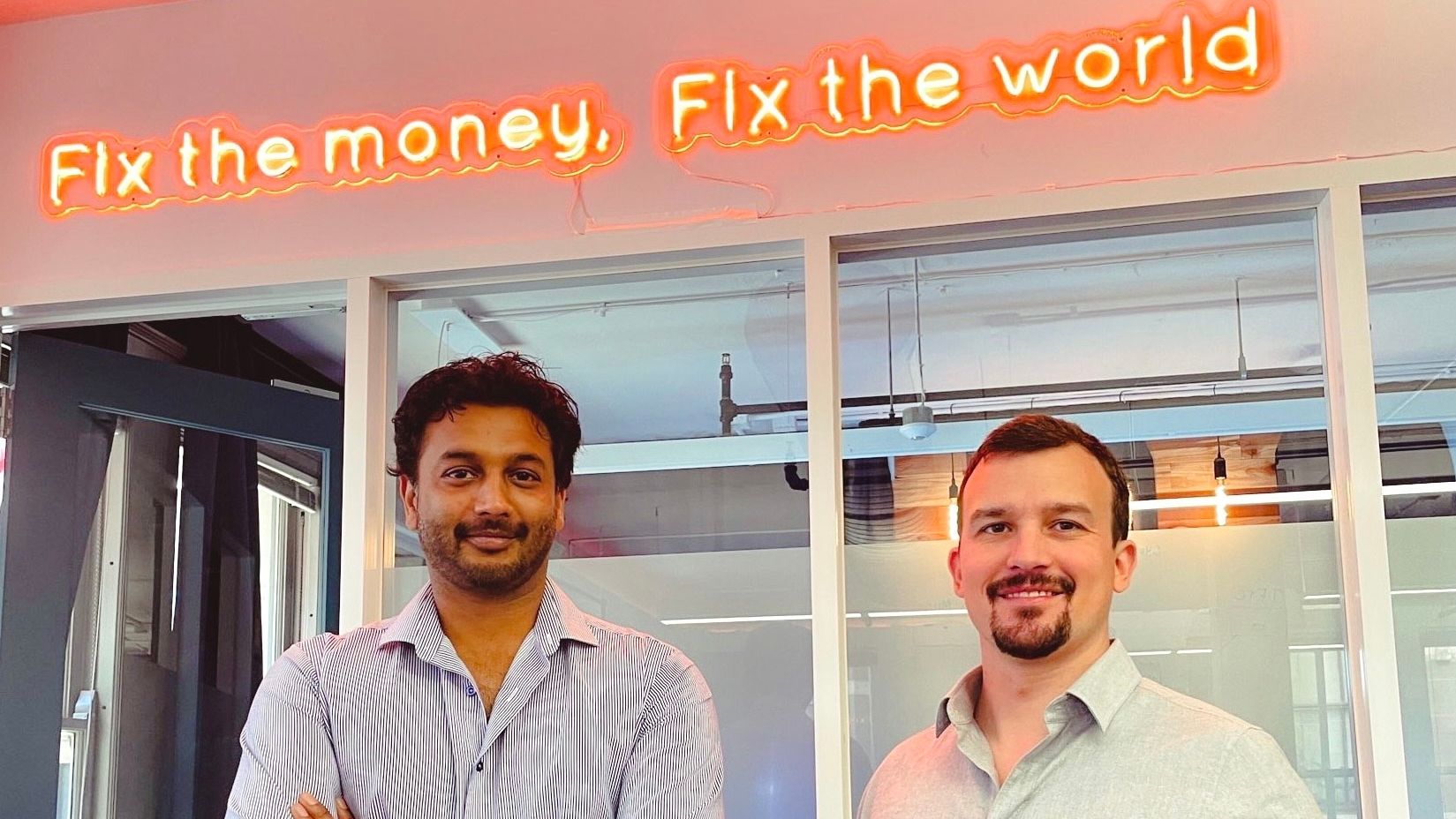 Unchained co-founders Dhruv Bansal and Joseph Kelly standing beneath a neon sign reading Fix the money, Fix the world