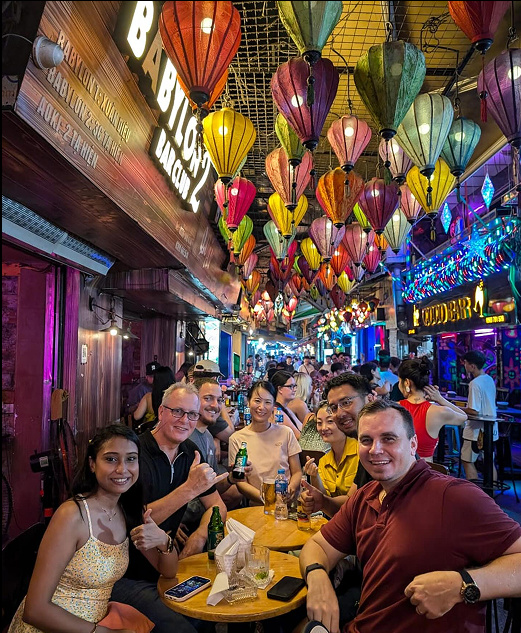 Joseph Ricard seated at the front right of a restaurant table with a group beneath colorful lanterns