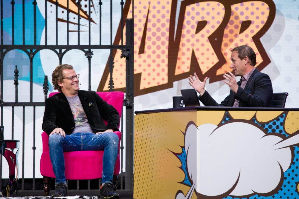 Josh James, seated left in a pink chair, speaking with Jason Lemkin on a colorful SaaStr conference stage.