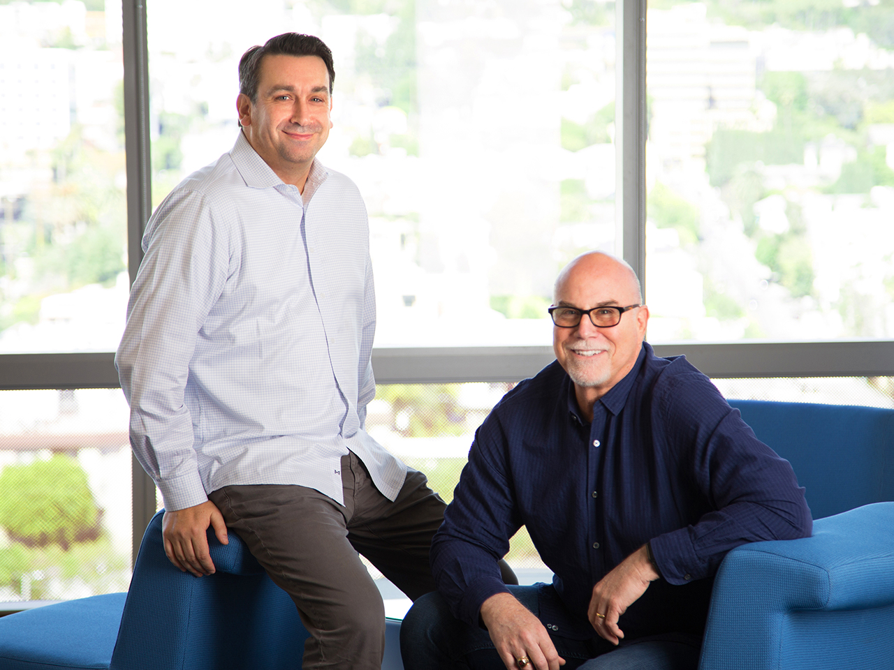 Josh Millet standing beside David Sherman, who is seated on a blue sofa