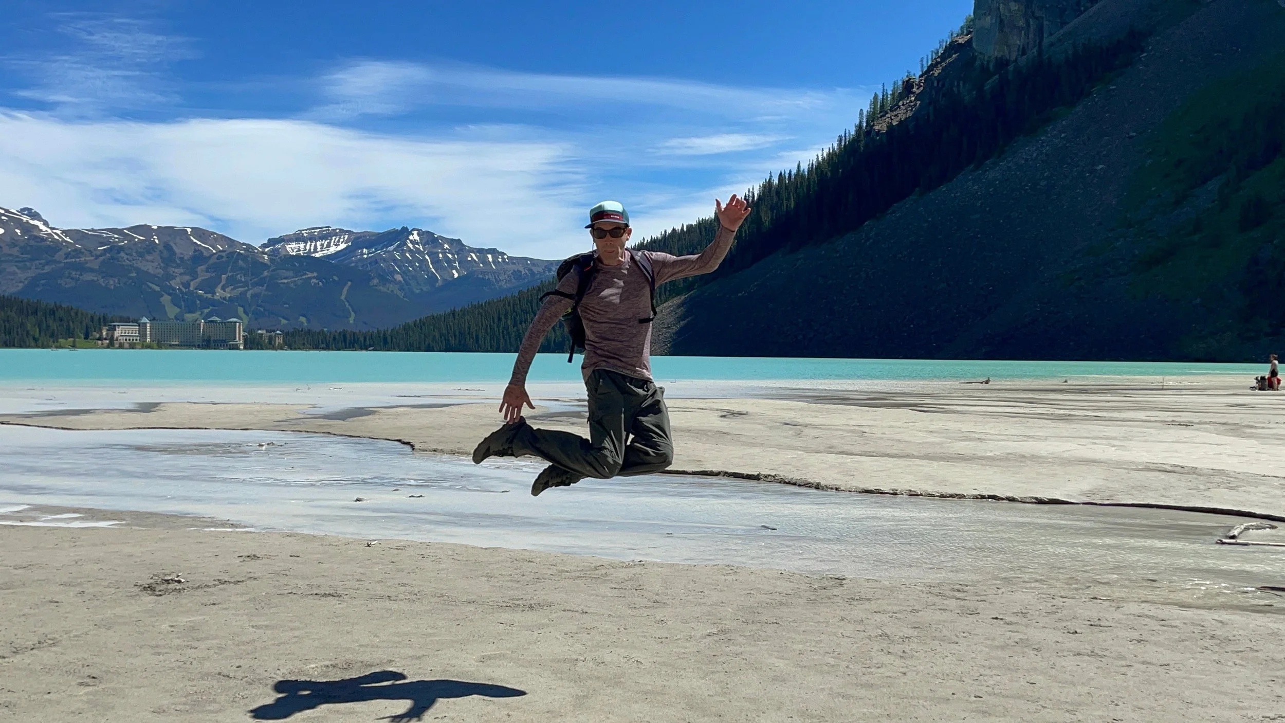 Joshua Dorkin jumping beside a turquoise lake with mountains behind him