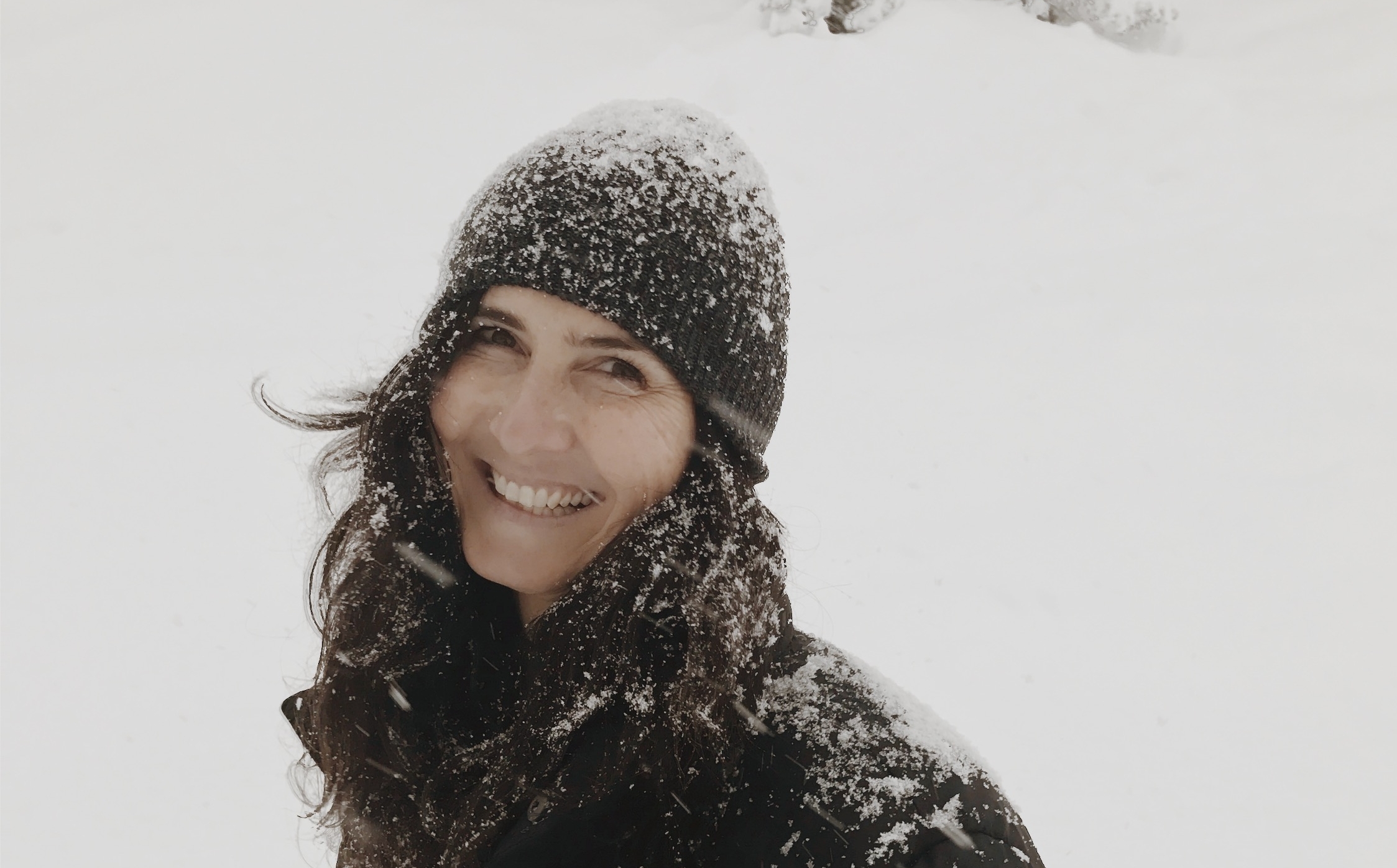 Joy Busse smiling outdoors in falling snow, wearing a dark knitted hat