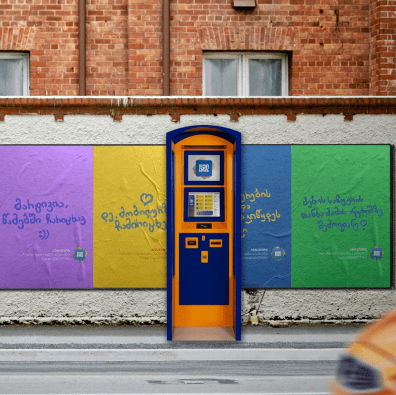 A blue and yellow PayBox payment terminal against a wall of colorful campaign posters