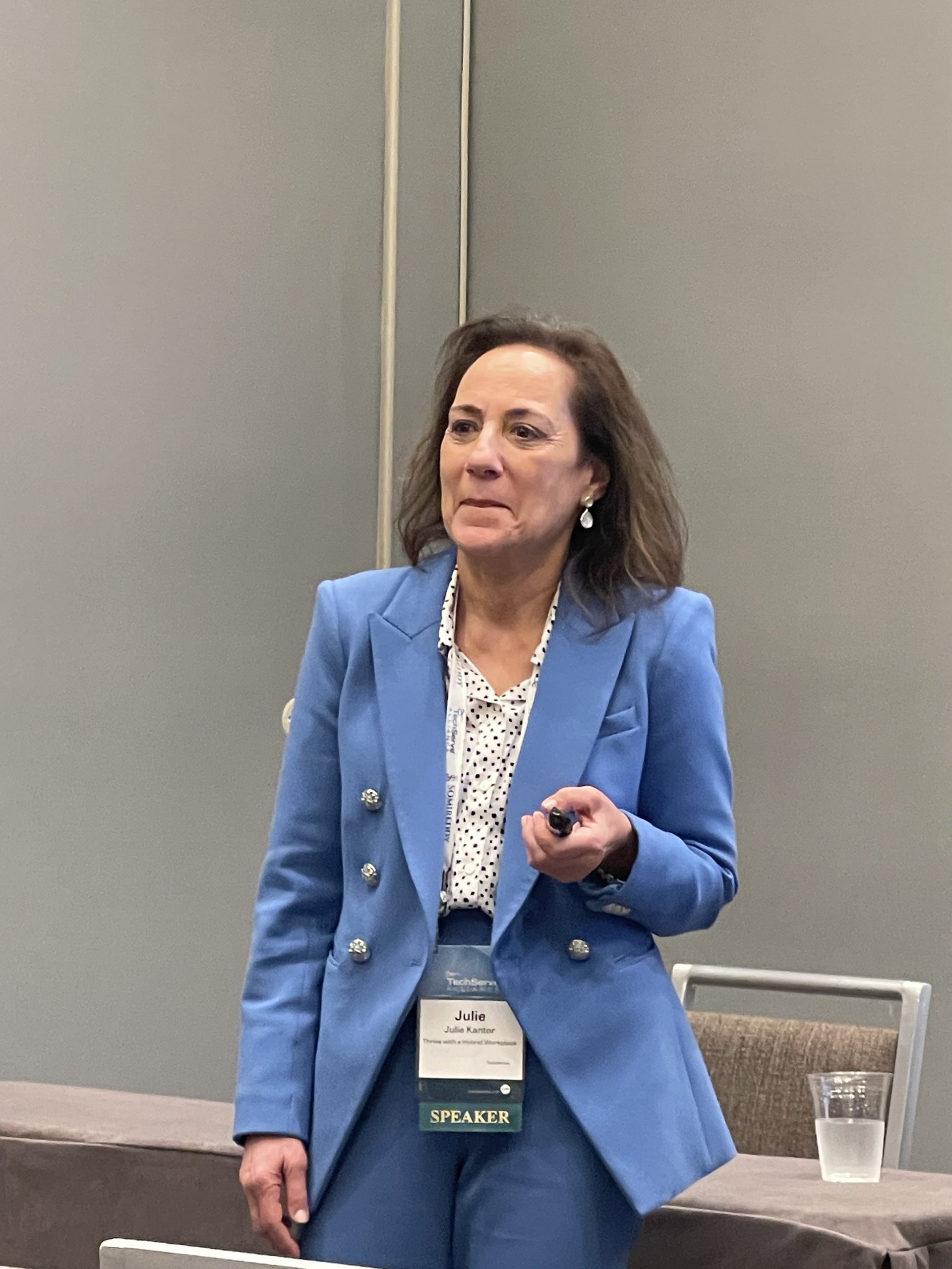 Julie Kantor speaking at a professional event in a blue suit, holding a presentation clicker