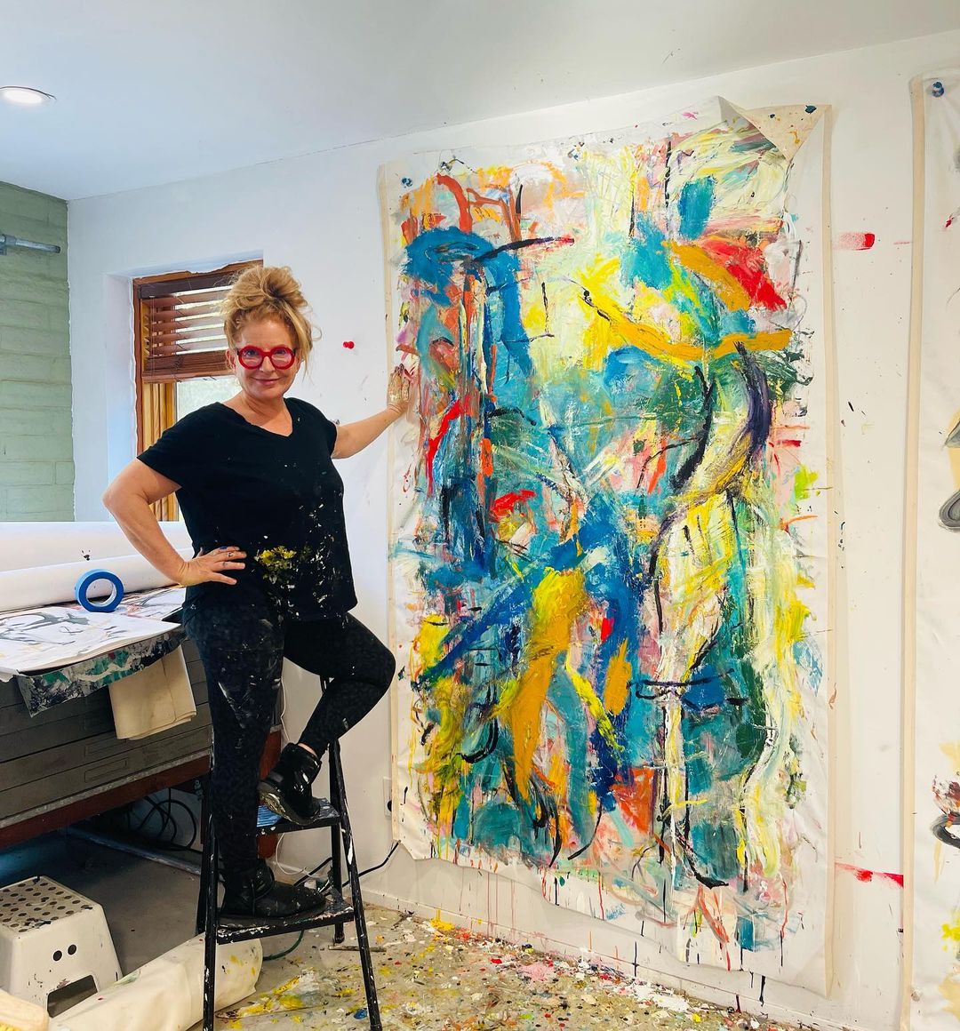 Julie Markfield wearing red glasses and paint-spattered clothes beside a large abstract painting in her studio