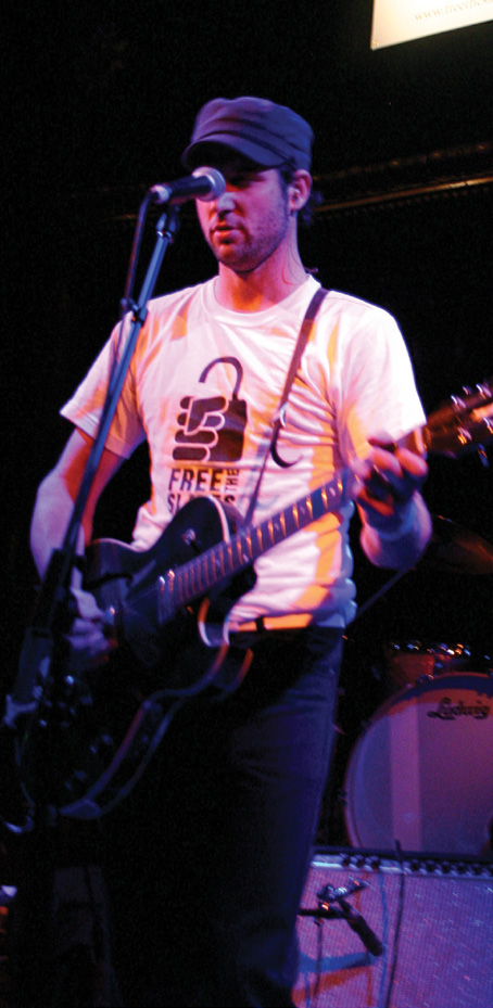 Justin Dillon playing electric guitar and singing onstage in a cap and Free the Slaves shirt