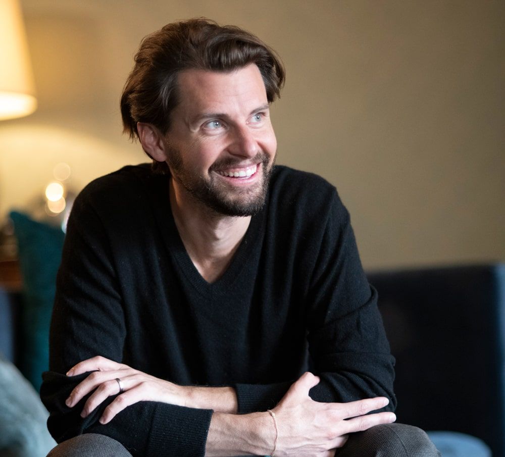 Justin McLeod seated on a blue sofa, smiling during a Hinge press photograph
