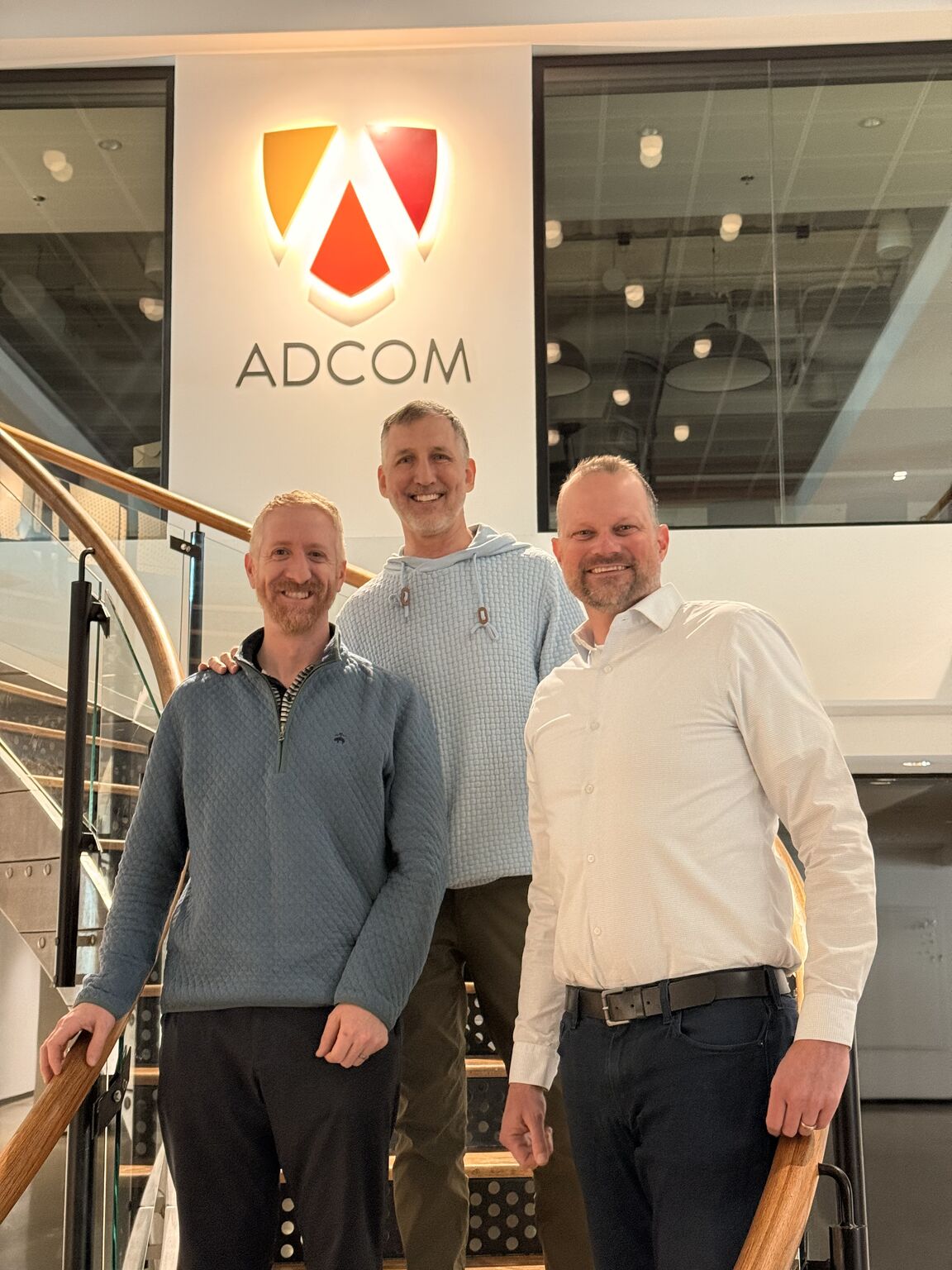 Three Adcom colleagues standing together on the office staircase in the photograph accompanying the co-CEO announcement