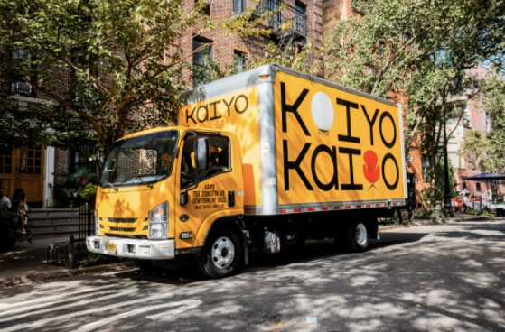 Yellow Kaiyo furniture delivery truck parked on a tree-lined city street