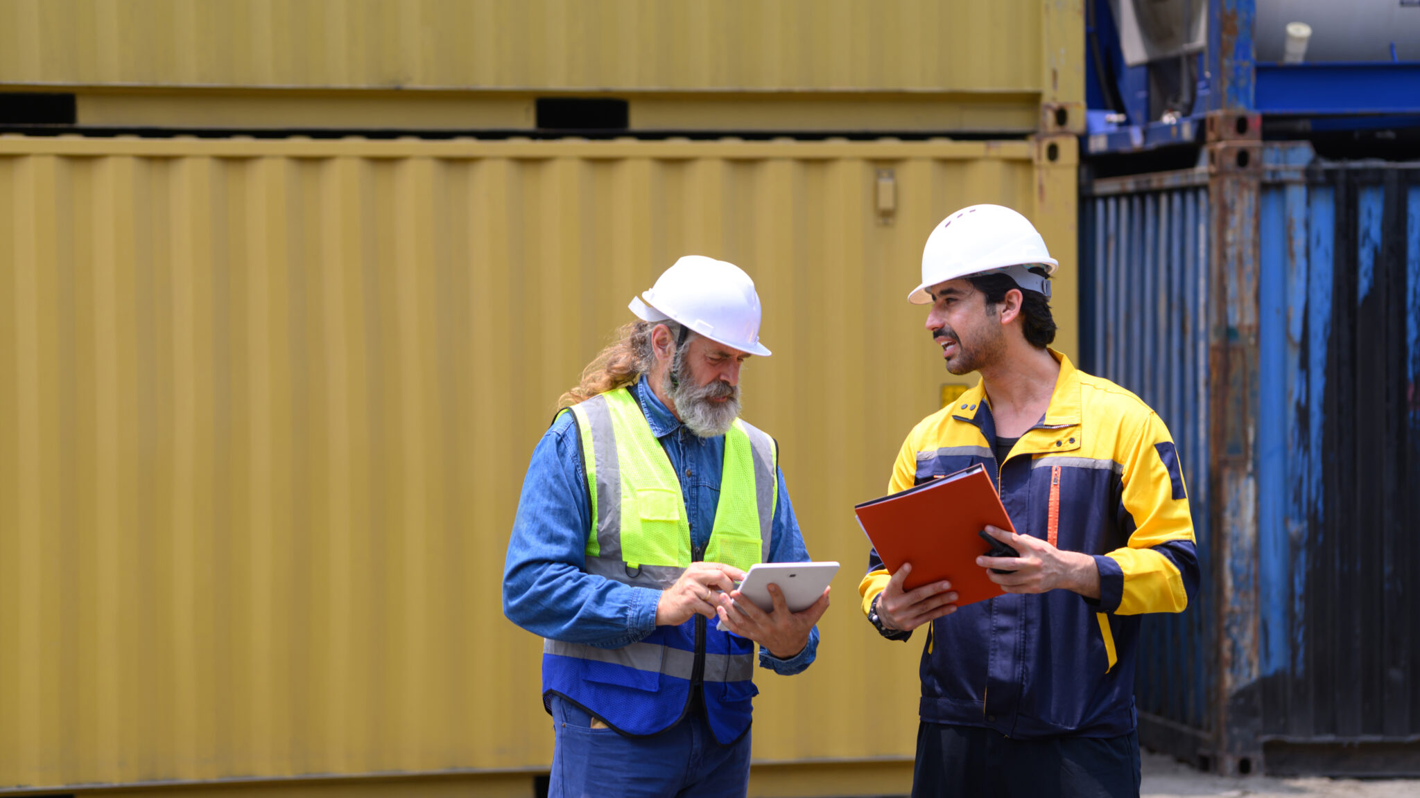 Two logistics workers using a tablet beside stacked shipping containers
