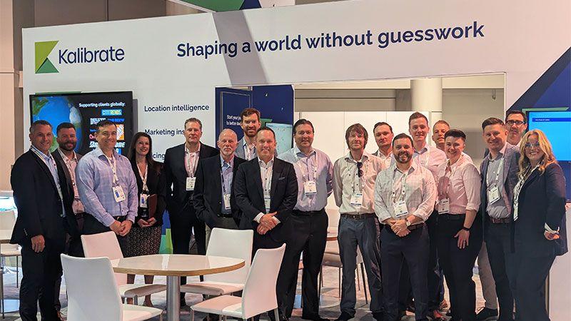 Kalibrate team gathered at a trade show booth beneath a sign about a world without guesswork