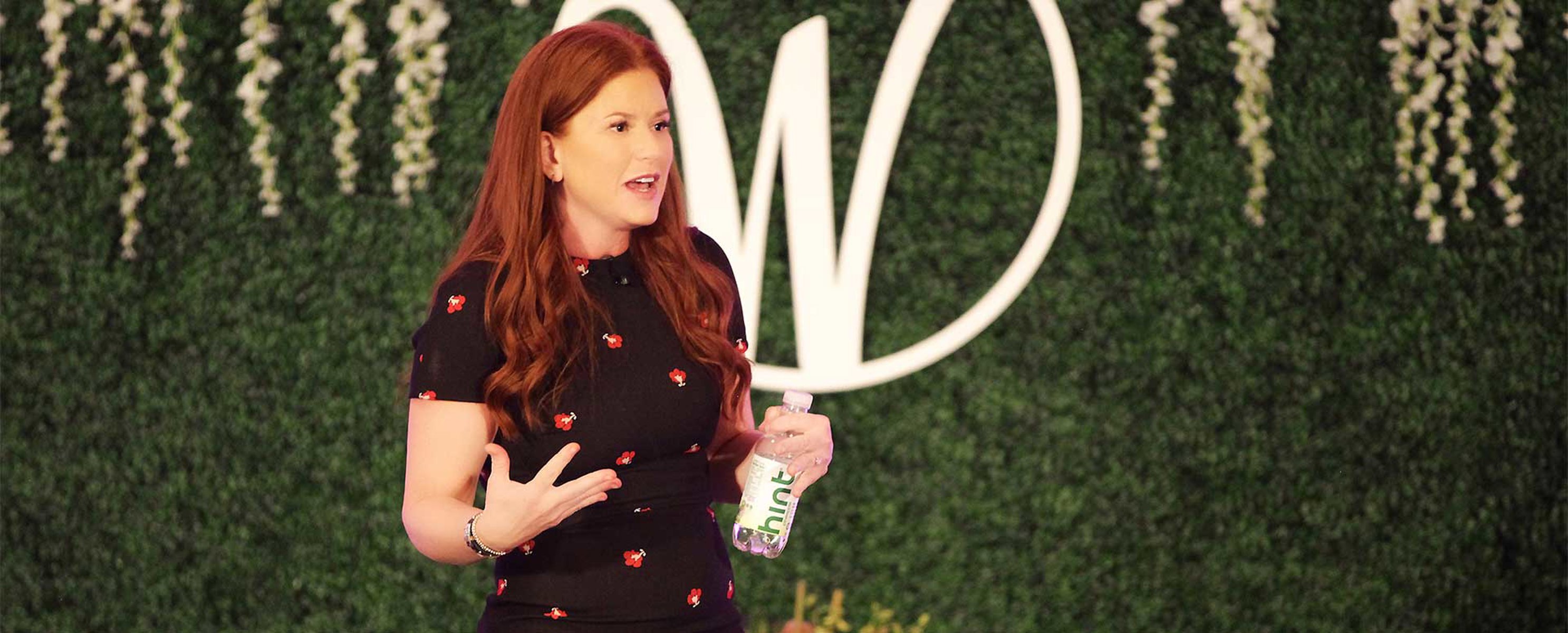 Kara Goldin speaking onstage while holding a bottle of Hint water