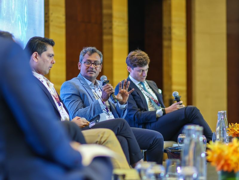 Karthik Murugian holding a microphone while speaking on the BIIA 2025 panel.