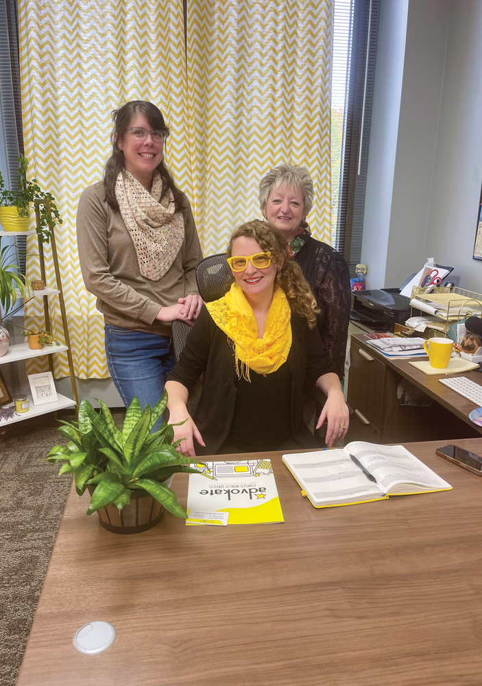 Kate Austin seated at a desk in yellow glasses and a yellow scarf, with two Advokate colleagues behind her