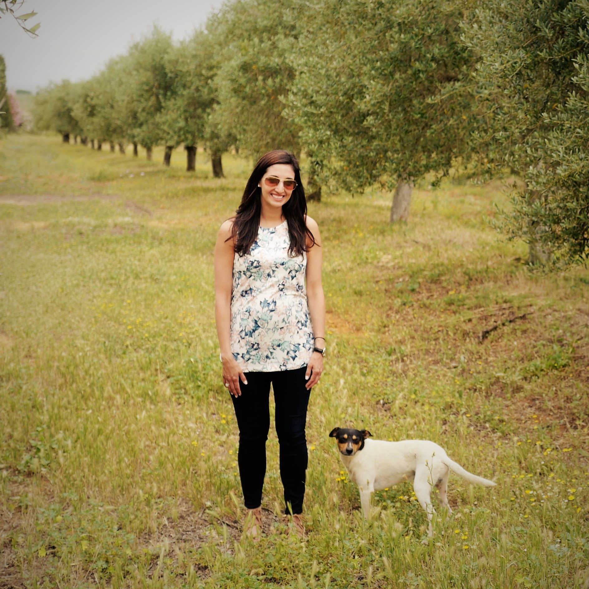 Katina Mountanos standing among olive trees in Greece, with a dog beside her