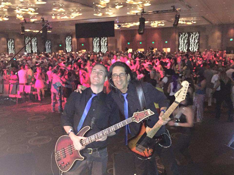 Kevin Ames, left, holding a bass guitar beside a fellow musician at a crowded event