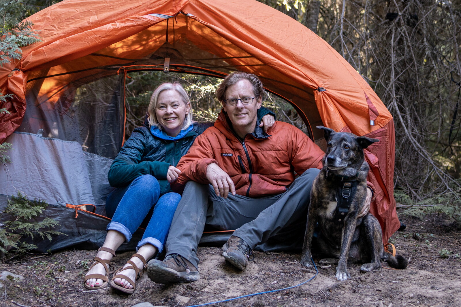 Sarah Smith and Kevin Long sitting with their dog Brandy at the entrance to an orange tent in the woods
