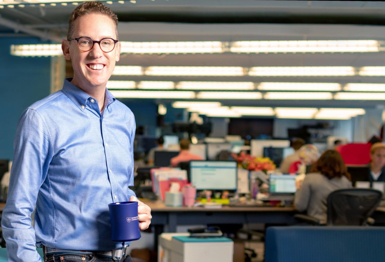 Kevin MacDonald holding a blue mug in Kit Check’s open-plan office, with colleagues working behind him