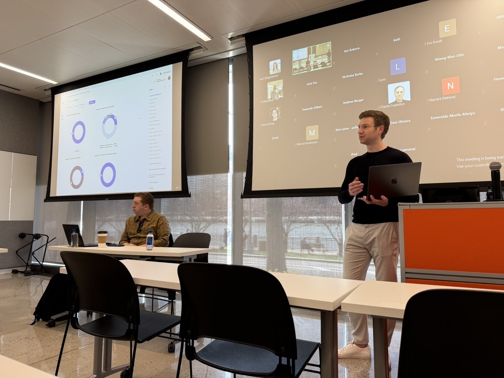 Kevin Walker holding a laptop while presenting alongside projected charts and remote meeting participants