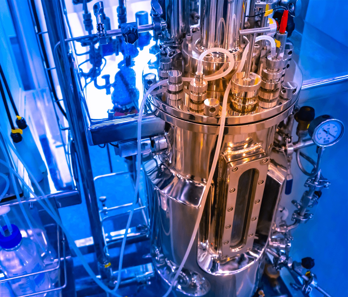 Stainless steel bioreactor with tubing and gauges under blue laboratory lighting