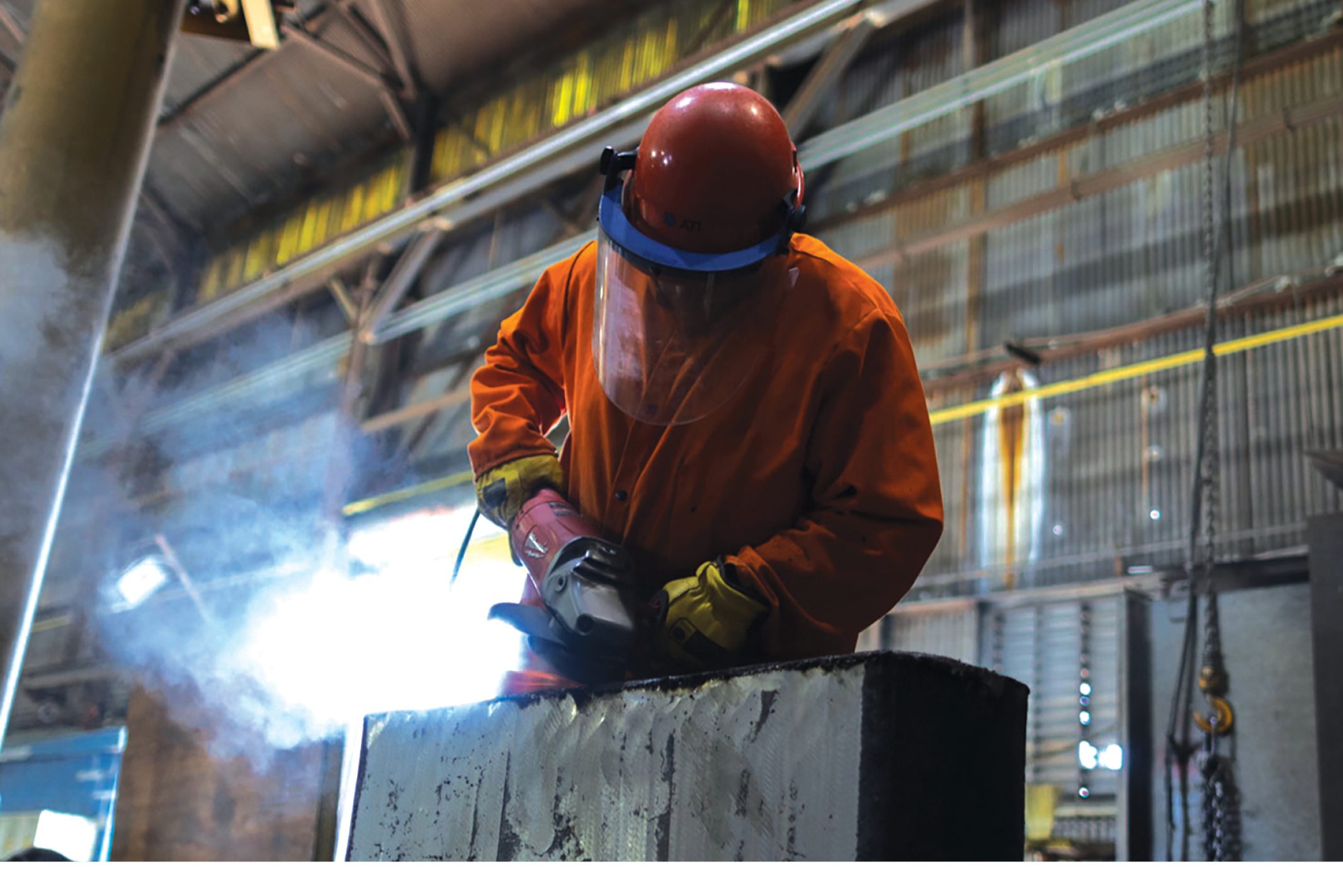 A worker in an orange protective coat and face shield uses a power tool on metal inside an industrial building