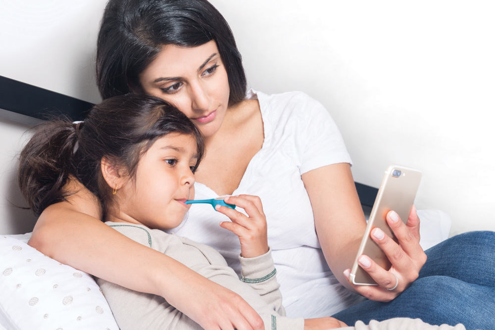 A parent checks a child's temperature with a Kinsa thermometer while viewing the companion phone app