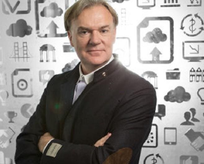 Archival press photograph of Kirk Krappe standing with folded arms against a background of business and cloud icons