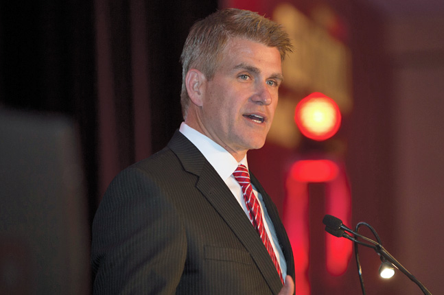 Kirk Perry speaking at a microphone, wearing a dark suit and striped tie