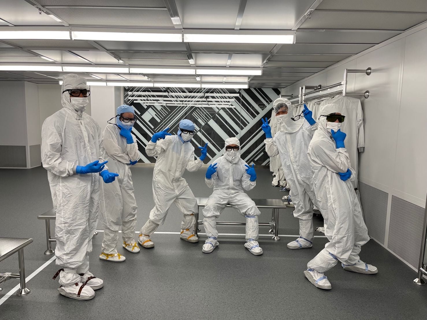 Six KLA team members in cleanroom suits, goggles and blue gloves posing together