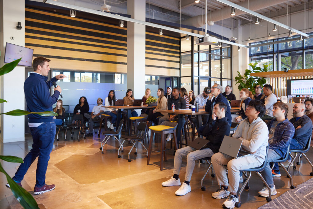 Koddi employees listen to a speaker during an office meeting, with laptops at communal tables