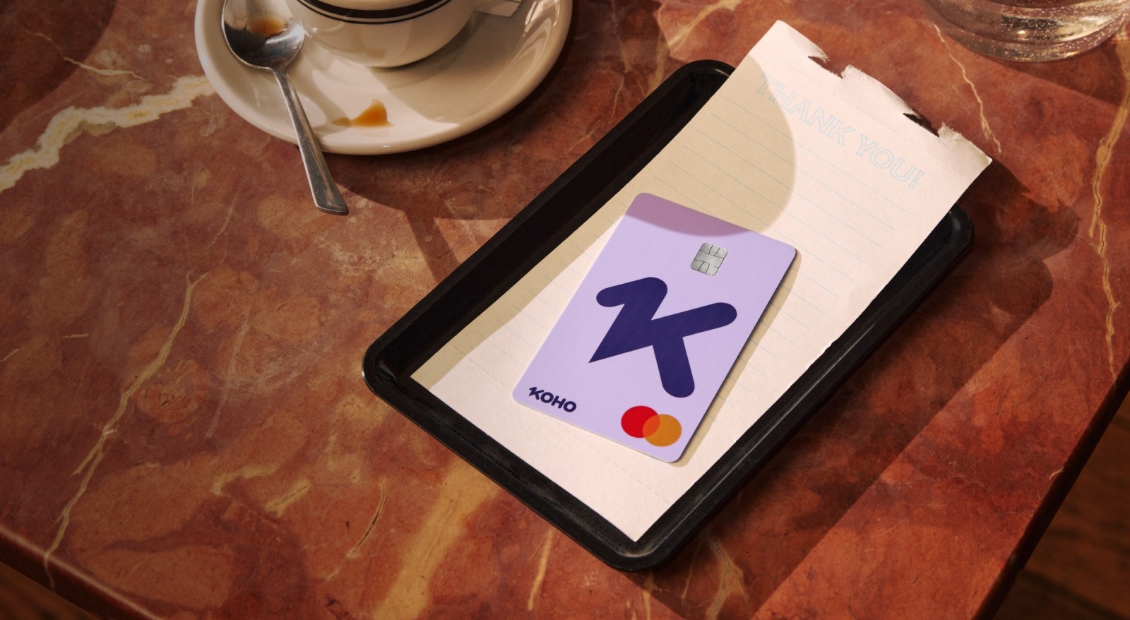A lavender KOHO prepaid Mastercard rests on a café receipt beside a coffee cup.