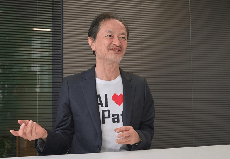Koichi Hasegawa gesturing during an interview, wearing a jacket over a UiPath T-shirt