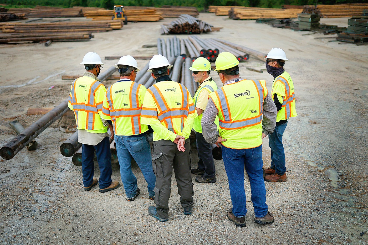 Six Koppers employees in reflective vests and hard hats standing among utility poles