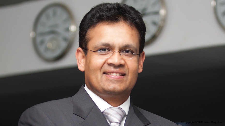 Kris Canekeratne in an office setting, with clocks on the wall behind him
