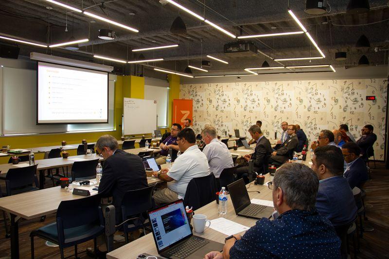 Forum attendees seated at tables with laptops, facing a presentation screen