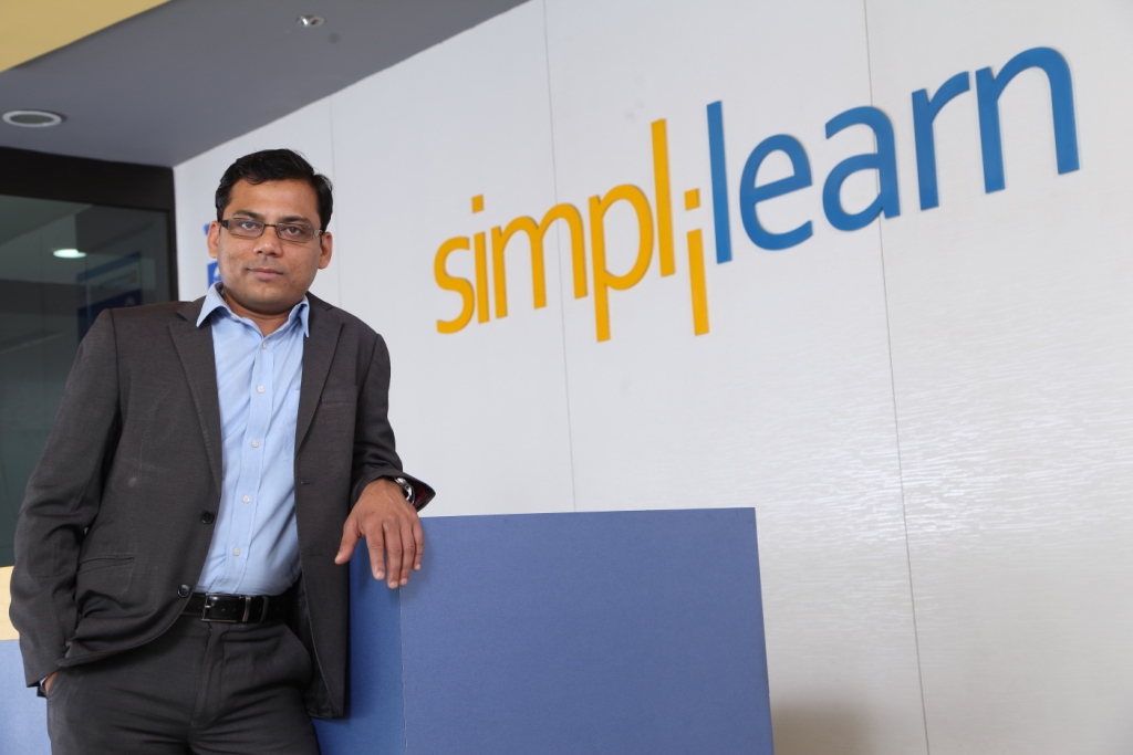 Krishna Kumar standing beside Simplilearn signage in an office