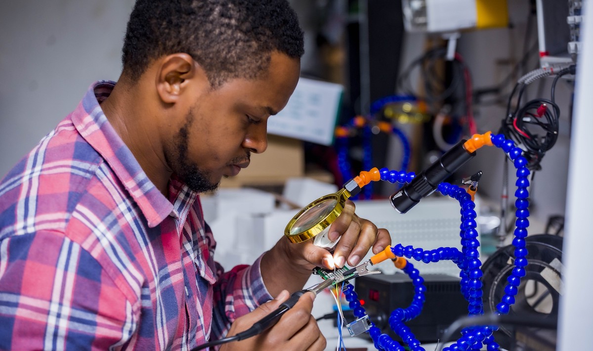 A maker at Kumasi Hive examines a circuit board under a magnifying glass while working with a soldering iron.