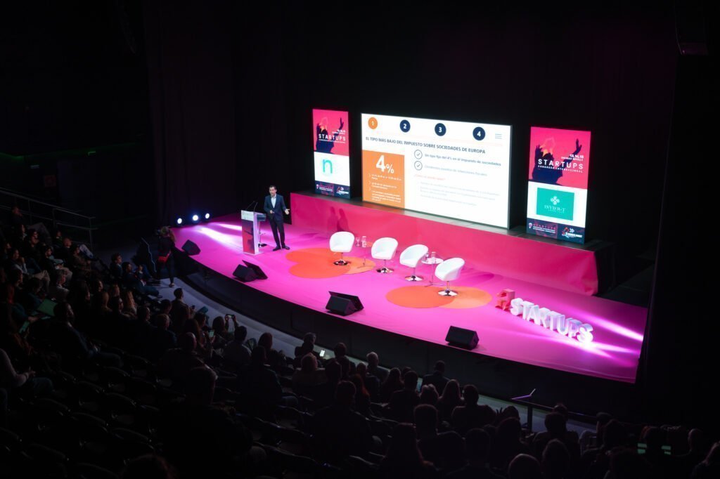A speaker addresses an auditorium from a brightly lit pink stage at the International Startup Congress