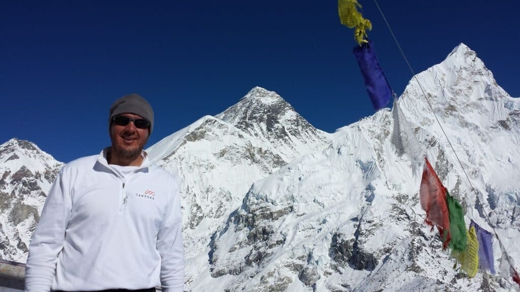 Lance Platt in a white jacket and sunglasses at Mount Everest base camp, with snowy mountains behind him