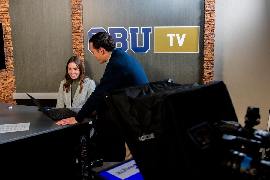 Two people working at the CBU TV anchor desk, with a studio camera in the foreground