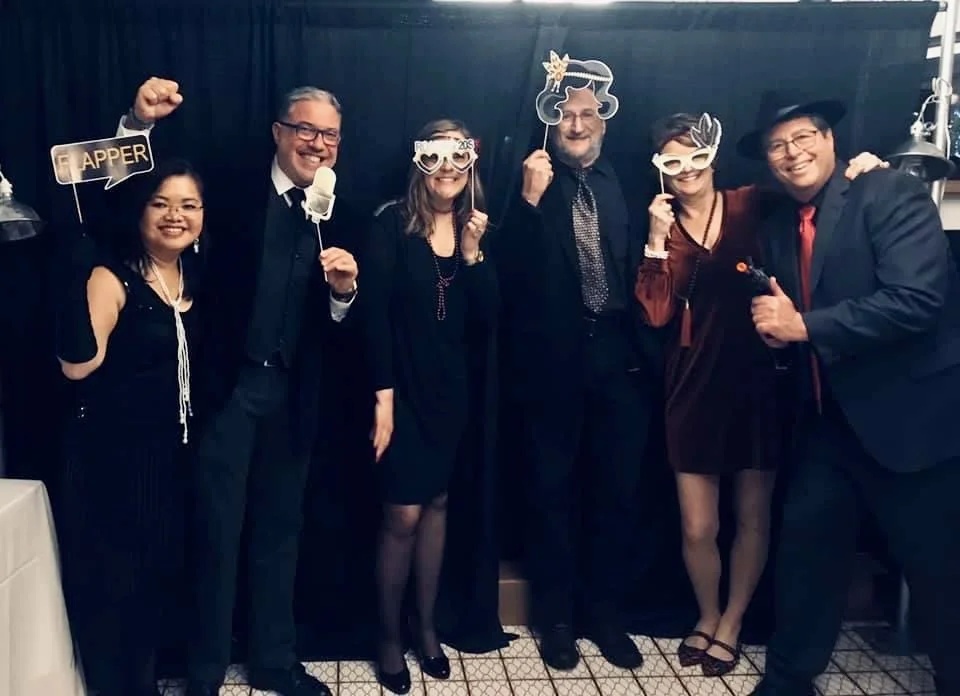Sage colleagues in party clothes posing with paper hats, masks and a mustache prop