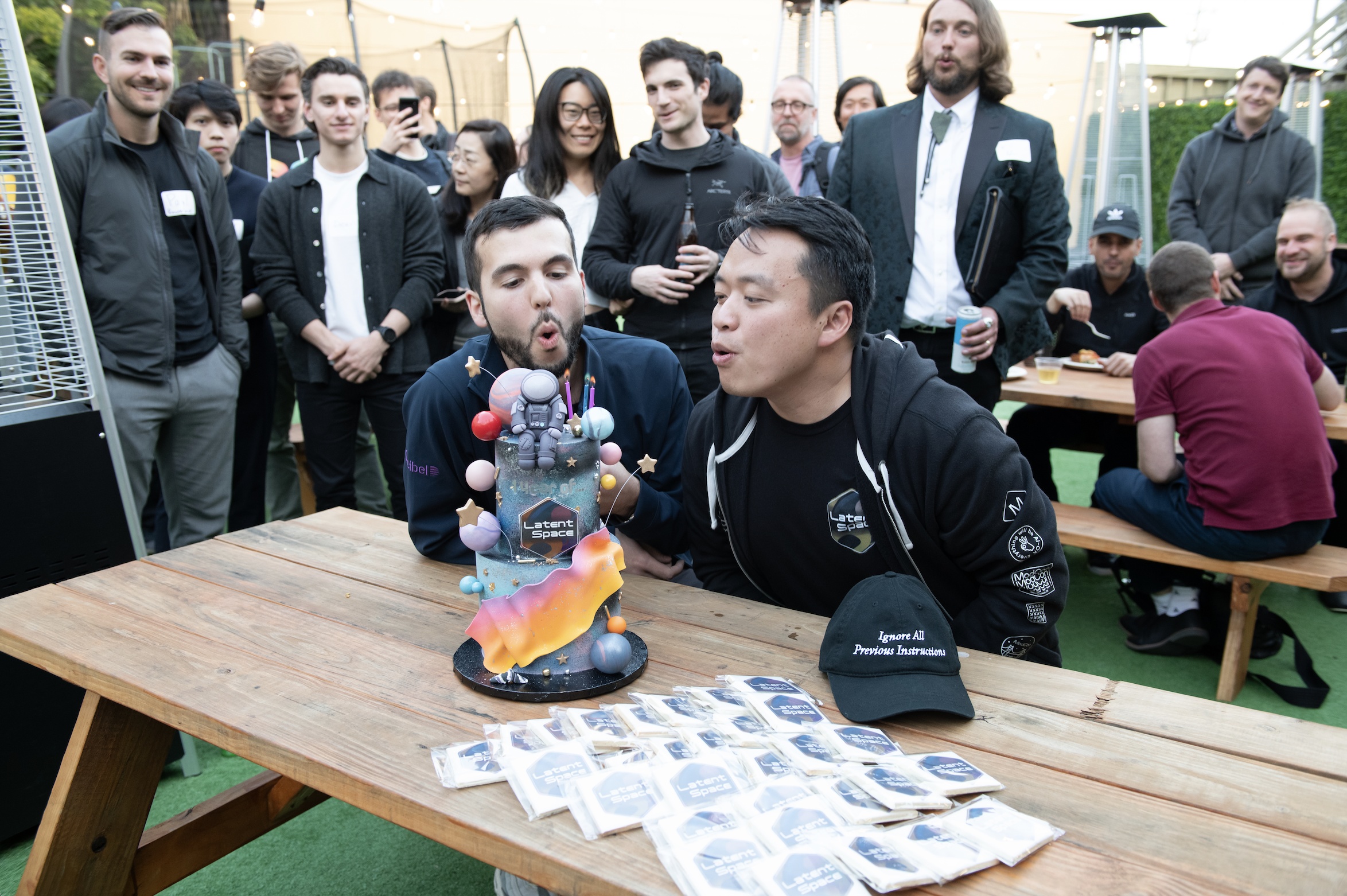 Alessio Fanelli and swyx blowing out candles on a space-themed birthday cake, surrounded by attendees